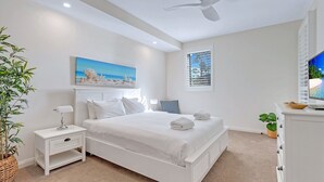 A bedroom with a bed, bedside tables, a ceiling fan, a painting, a window with blinds, and a television. - Le Maison at the Beach (Huskisson)