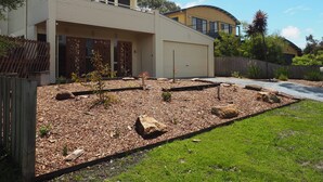 A modern two-story house with a garage, a well-maintained garden with mulch and rocks, and a wooden fence. - The Beachcomber , Linen Supplied, 10 minute Walk to Surf Beach (Inverloch)