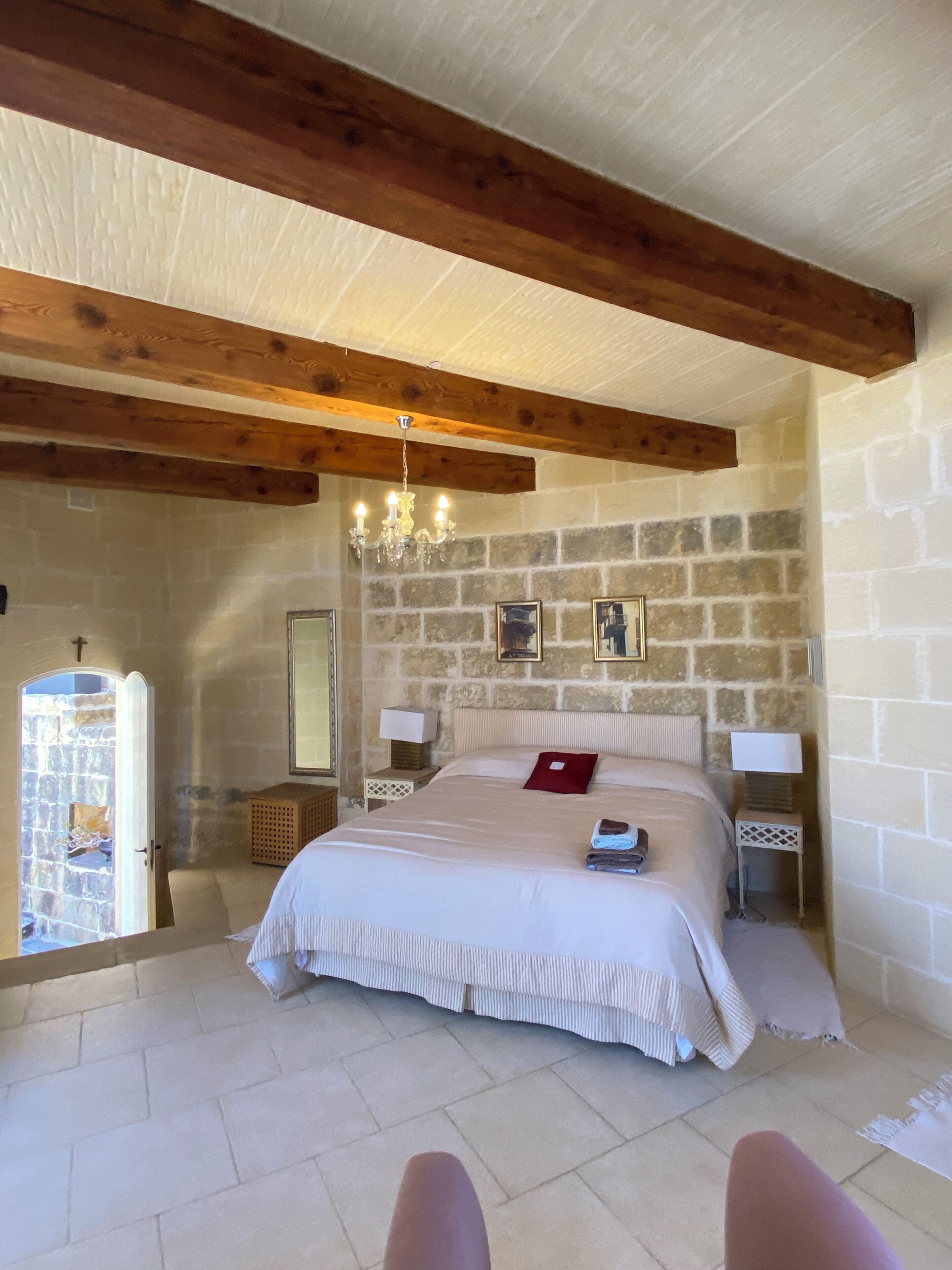 A bedroom with a stone wall, wooden beams, a chandelier, a bed with a red pillow, and a nightstand with a lamp.