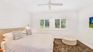 A bedroom with a bed, two windows with blinds, a ceiling fan, a small round ottoman, and a painting on the wall.