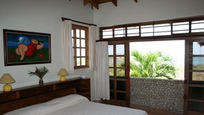 Un dormitorio con cama, dos lámparas de noche, una mesita de noche, un cuadro, una ventana con cortinas y vistas a zonas verdes a través de un ventanal.