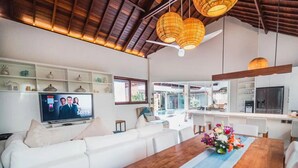 A modern living room with a white sofa, a wooden dining table with a blue runner, and wicker pendant lights. A TV is mounted on the wall displaying a show.