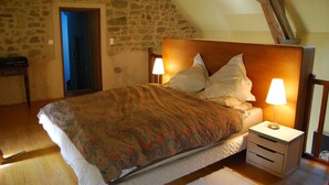 Ein Schlafzimmer mit Steinwand, Holzbalkendecke, einem Bett mit gemustertem Überwurf, zwei Nachttischlampen und einem Nachttisch mit Schubladen.