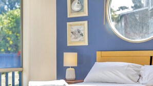Un lit bien fait, avec du linge de lit blanc, une fenêtre ronde et deux tableaux encadrés accrochés au mur.