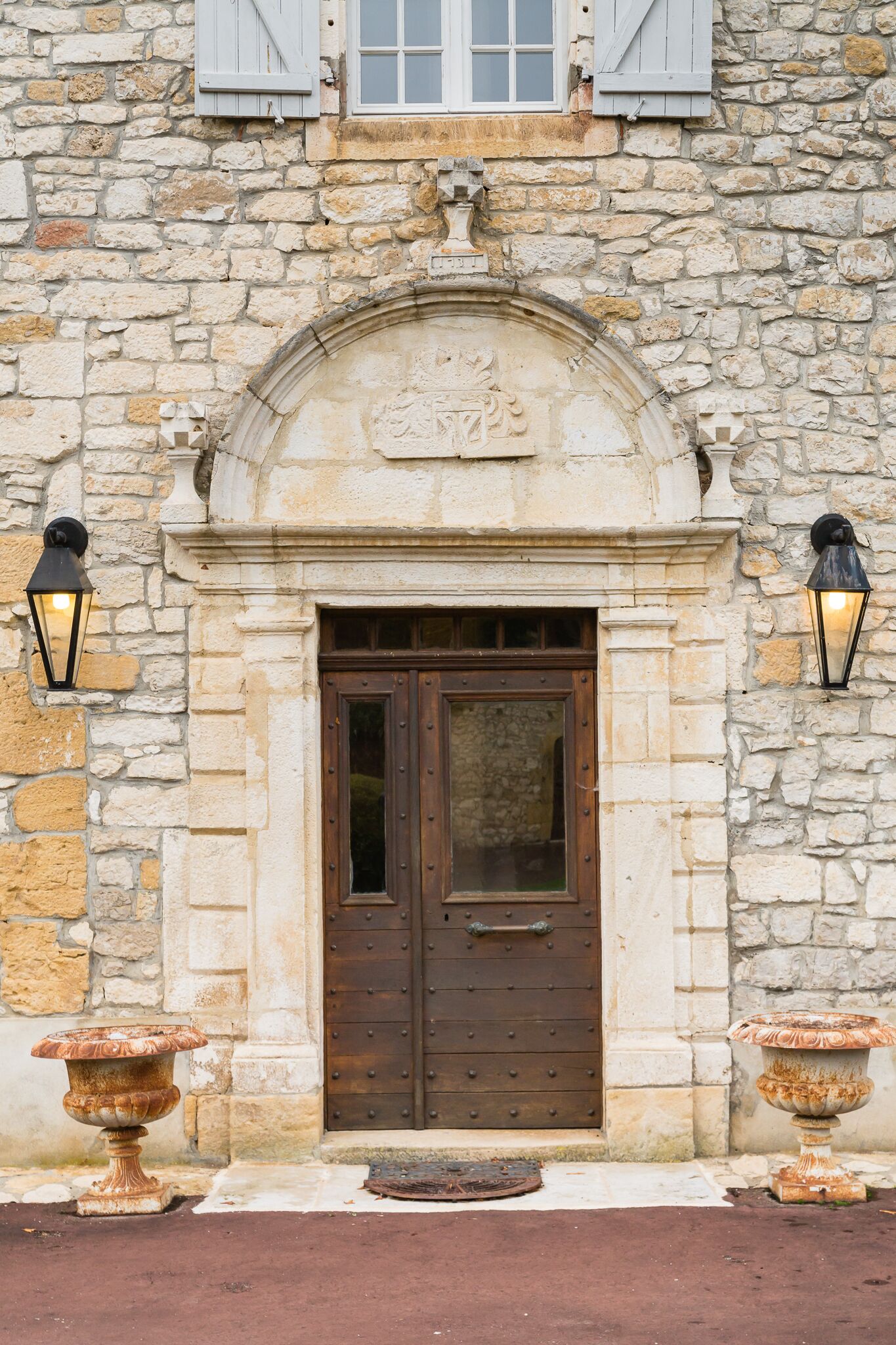 A stone building with a wooden door, two wall-mounted lanterns, and a decorative urn on either side of the door.