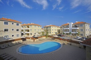 A hotel with a swimming pool surrounded by sun loungers and a few cars parked nearby.