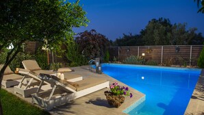 A swimming pool with a wooden deck, lounge chairs, and a rock planter with flowers. - Villa Komfort in Zakynthos suitable for family or group with private pool (Lagopodo,Zakynthos)