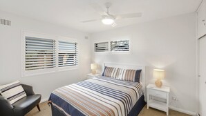 Une chambre à coucher comprenant un lit, deux tables de chevet, une chaise, un ventilateur de plafond et une fenêtre avec des stores.