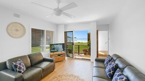 Un salon avec deux canapés en cuir, un ventilateur de plafond et un téléviseur posé sur un meuble en bois. On y trouve une grande fenêtre avec des stores, une porte donnant sur un balcon avec vue sur la plage, et une petite horloge ronde accrochée au mur.