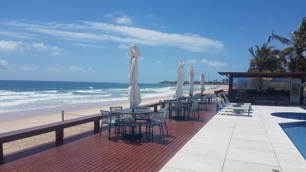 Une terrasse en bois avec coin salon extérieur et espace piscine, offrant une vue sur la plage et l’océan.