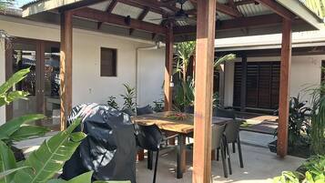 Une terrasse extérieure avec une table et des chaises en bois, entourée de plantes et dotée d’un espace abrité avec un toit en métal.