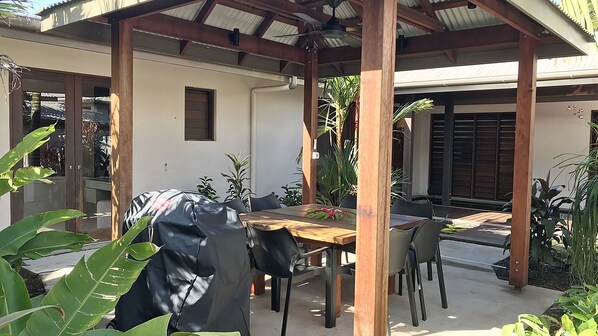 Une terrasse extérieure avec une table et des chaises en bois, entourée de plantes et dotée d’un espace abrité avec un toit en métal.
