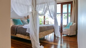 A bedroom with a four-poster bed, a sitting area with a chair, and a view of the outdoors.