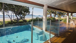 Une piscine à l’eau d’un bleu cristallin, entourée d’une terrasse en bois et d’une balustrade en verre, offrant une vue sur une plage et des montagnes.