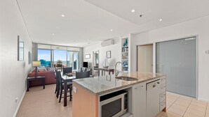 A modern kitchen with a built-in dishwasher, a dining area with a table and chairs, and a view of the ocean through large windows. - The Penthouse with Views (Victor Harbor)