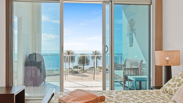 A hotel room with a large sliding glass door leading to a balcony with a view of the ocean and palm trees. - The Penthouse with Views (Victor Harbor)