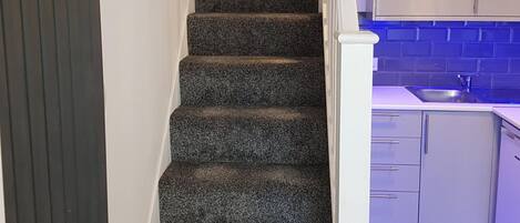 A staircase with dark carpeted steps leading to a kitchen area with white cabinets and a blue-lit countertop.