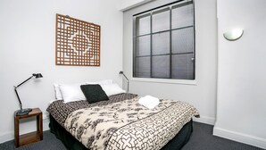 A bedroom with a bed, a nightstand, a lamp, a window with blinds, and a decorative screen. - Sydney- CLD01 Bridge Street - Modern 1BR with Rooftop Pool in the Heart of Sydney CBD (Sydney)