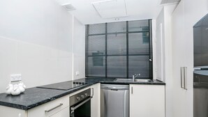 A compact kitchen with white cabinets, a black countertop, a built-in oven, a dishwasher, and a window with blinds. - Sydney- CLD01 Bridge Street - Modern 1BR with Rooftop Pool in the Heart of Sydney CBD (Sydney)