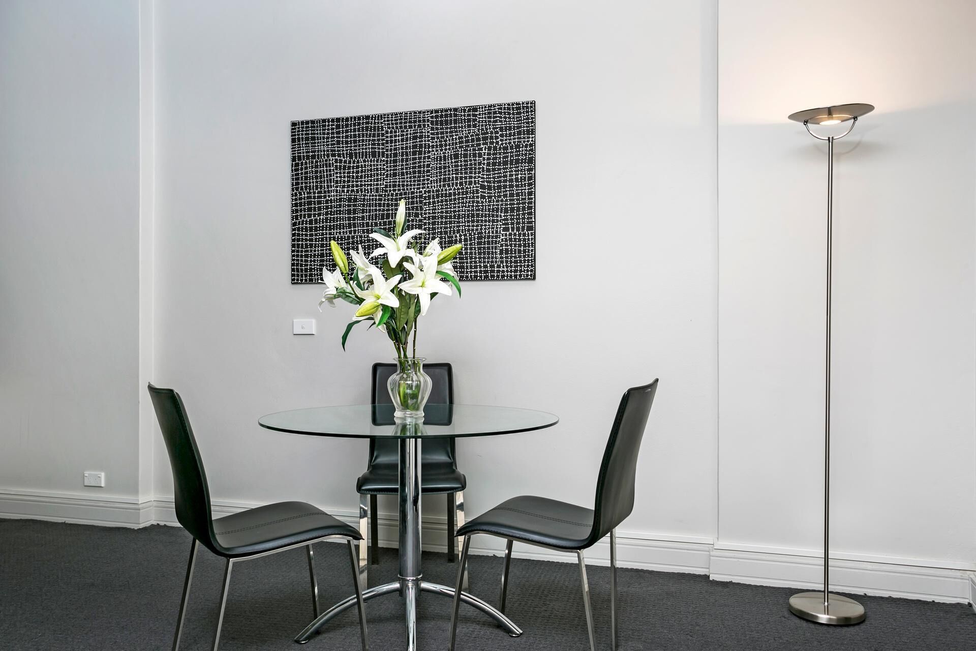 A modern dining area with a glass table, two black chairs, a vase with white flowers, and a floor lamp.
