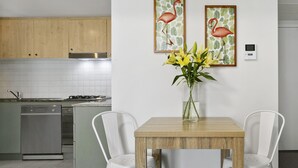 A modern kitchen with wooden cabinets, a stainless steel stove, and a white tiled backsplash. There is a dining table with two white chairs and a vase of yellow flowers on the wall.