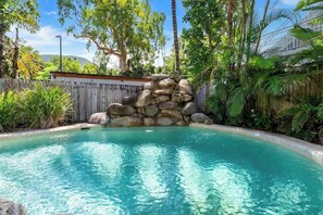 A swimming pool with clear water, surrounded by a wooden fence, palm trees, and a rocky structure.
