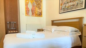 Un lit bien fait, avec du linge de lit blanc et des serviettes pliées, une tête de lit en bois et un tableau représentant Londres accroché au mur.