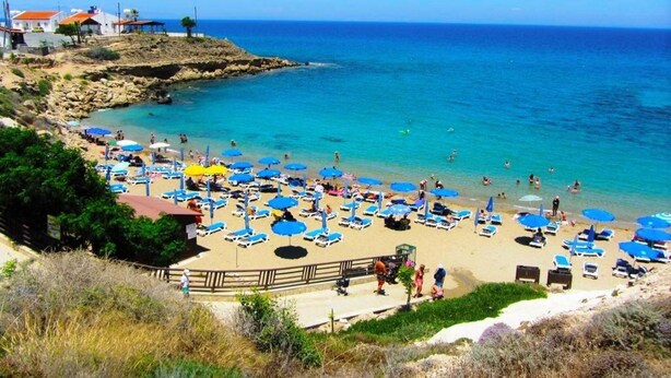 Une plage dotée de nombreuses chaises longues et de parasols, un rivage rocheux et une mer d’un bleu limpide.