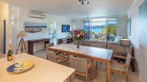 A spacious living area with a large window offering a view of the sea, a dining table with chairs, and a sofa. - Lagoon Lodge 105, Beachfront Apartment on Hamilton Island (Hamilton Island)