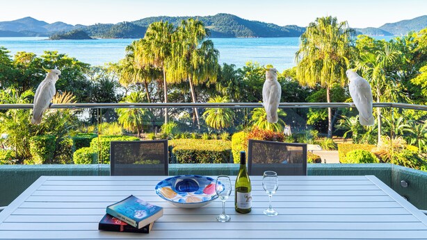 A balcony with a table set for two, overlooking a scenic view of a lake and mountains. - Lagoon Lodge 105, Beachfront Apartment on Hamilton Island (Hamilton Island)