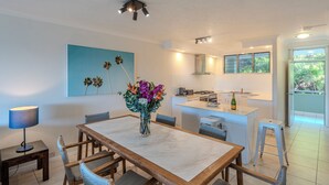 A modern kitchen with white cabinets, a central island, and a dining area with a marble table and wicker chairs. - Lagoon Lodge 105, Beachfront Apartment on Hamilton Island (Hamilton Island)
