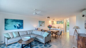 A modern living room with a sectional sofa, a dining area with a wooden table, and a kitchen in the background. - Lagoon Lodge 105, Beachfront Apartment on Hamilton Island (Hamilton Island)