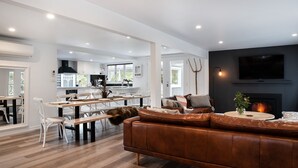 A modern living room with a leather sofa, a dining area with white chairs, and a kitchen in the background.
