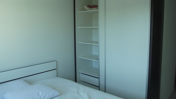 Een slaapkamer met een bed, een wit hoofdeinde, een garderobe met veel planken en een deur.