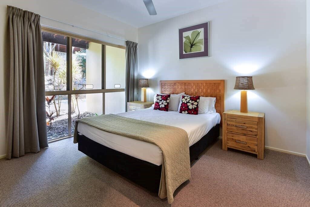 Quarto de hotel com uma cama grande, duas mesas de cabeceira com luminárias, uma janela com cortinas e um quadro na parede.