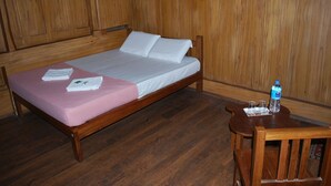 A wooden bed with a pink bedspread and white linens, a wooden nightstand with a water bottle and glasses, and a wooden chair. - Nature Castle-A traditional kerala style  (Vattavada)
