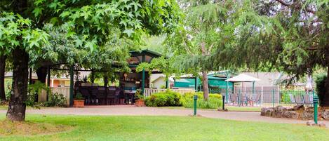 A covered outdoor seating area with tables and chairs, surrounded by lush greenery and trees.