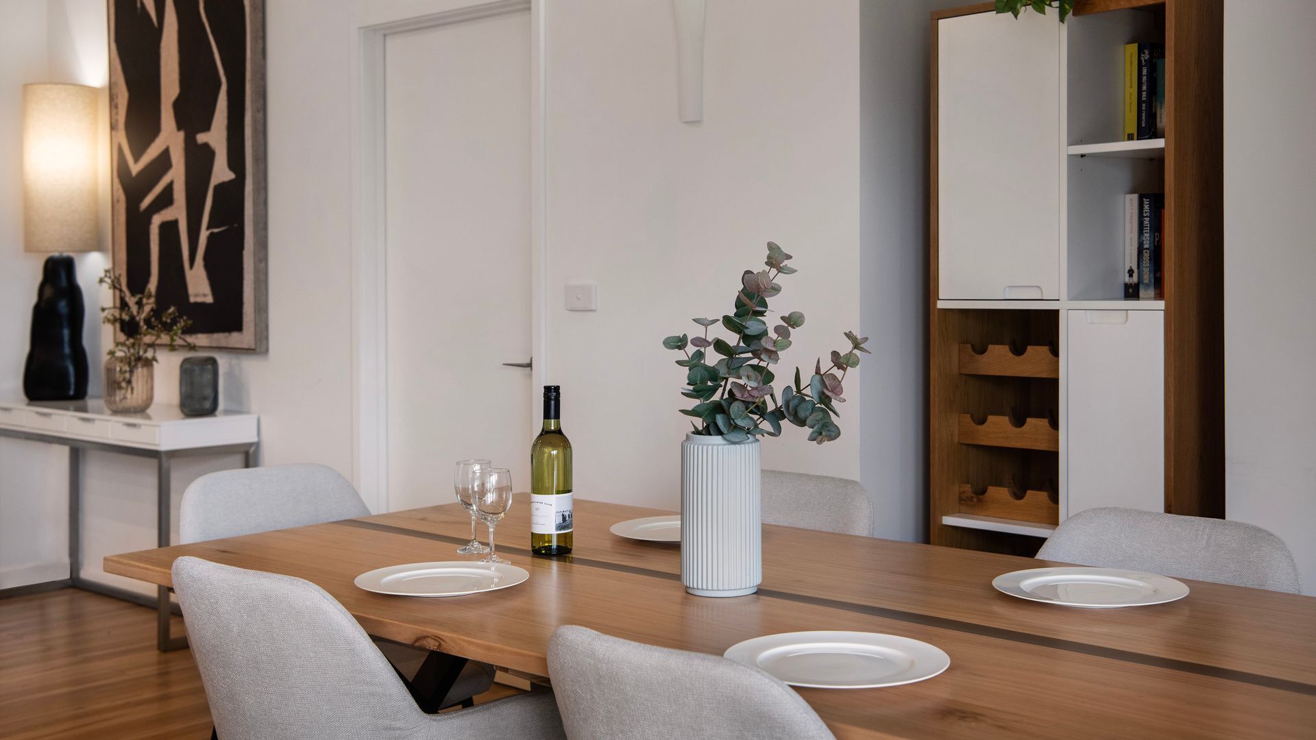 A dining area with a wooden table, four chairs, a wine bottle, and a vase with plants.