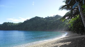 A beach with clear turquoise water, sandy shore, and lush green hills in the background. - Secluded Hillside Cottage with Bay & Rainforest Views, Just 5 Mins to the Beach (Englishman's Bay)