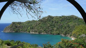 A coastal view through a circular window, featuring a lush green hillside, turquoise sea, and a clear sky. - Secluded Hillside Cottage with Bay & Rainforest Views, Just 5 Mins to the Beach (Englishman's Bay)