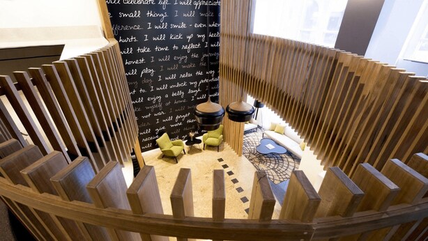 A wooden spiral staircase leading to a seating area with a blackboard with handwritten notes, green chairs, and a small table with a lamp.