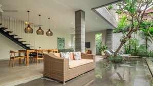 A modern living area with a wicker sofa, wooden chairs, a staircase, and a view of a lush garden. - Villa Jekumah 3 - 1 minute walk to restaurants (Kerobokan)