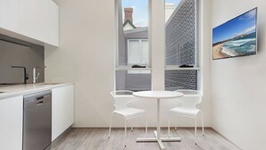 Una cucina moderna con mobili bianchi, lavastoviglie e una finestra con vista su un edificio. È presente un tavolo rotondo con due sedie bianche e un quadro raffigurante una spiaggia sulla parete.