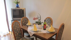 Ein Esstisch für vier Personen, mit Stühlen, ein Fernseher im Hintergrund und ein Blumentopf auf der Tischplatte.