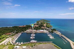 An aerial view of a marina with boats docked, a small island with buildings, and a clear blue sky.