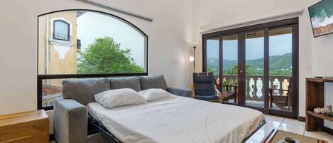 Un dormitorio con cama, sofá, sillón, mesita de madera, lámpara de pie y vistas a un balcón con paisaje panorámico.