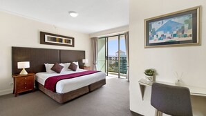 A hotel room with a large bed, a desk, a chair, a nightstand, a lamp, a painting, and a view of the outside.