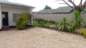 A residential property with a gravel driveway, a small garden with various plants, a concrete wall, and a building with a metal roof.