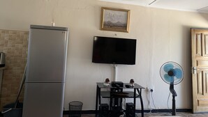 A compact living space with a refrigerator, a flat-screen TV mounted on the wall, a small table with electronic devices, and a standing fan.