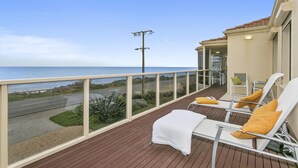 Een balkon met uitzicht op de oceaan, met twee witte lounge stoelen met gele kussens, een witte tafel en een klein zitgedeelte met een groene kussen.
